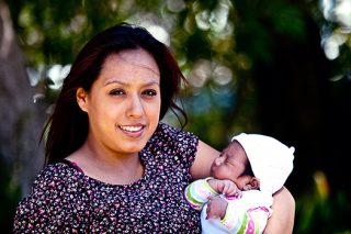 A photo of a young woman holding a newborn baby in her arms in a park while smiling at the camera.