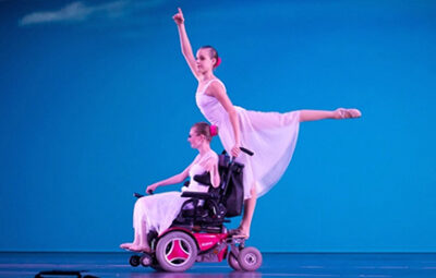 Jessica Clay in a motorized wheelchair wearing a flowing dancer costume, posed in an arabesque from the chair.