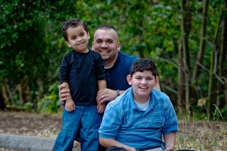 A portrait photo of a father and his two sons sitting near a park smiling at the camera.