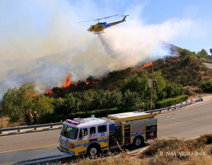 Picture of fire engine in foreground and background is a fire burning on the hill side sloping down with a helicopter dropping water on the fire